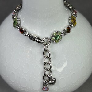 May include: Silver-toned bracelet featuring a floral design with colorful gemstones. The bracelet has a chain link style with small flower-shaped charms in green, yellow, and pink. A dangling charm with a pink gemstone hangs from the clasp.