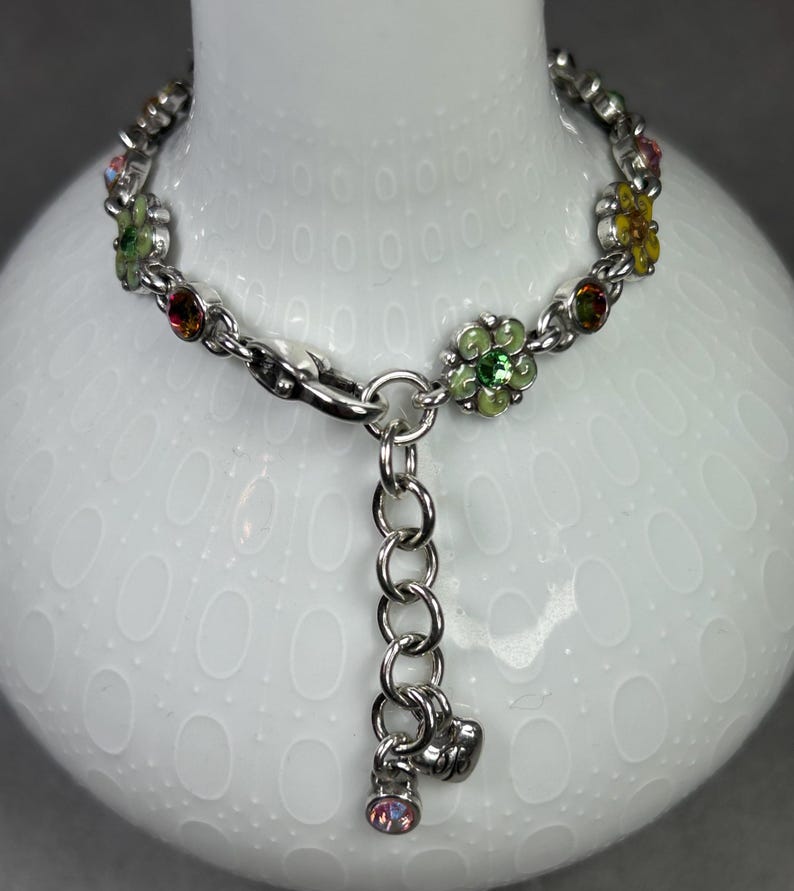 May include: Silver-toned bracelet featuring a floral design with colorful gemstones. The bracelet has a chain link style with small flower-shaped charms in green, yellow, and pink. A dangling charm with a pink gemstone hangs from the clasp.