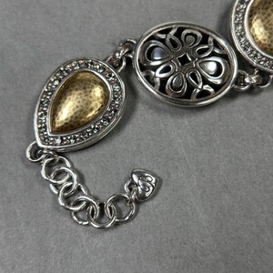 以下が含まれることがあります： 3つの装飾的なリンクを備えたシルバーカラーのブレスレット。リンクは、涙の形、花のパターン、円など、金と銀のアクセントがあります。ブレスレットは、小さな丸いリンクのチェーンで接続されています。