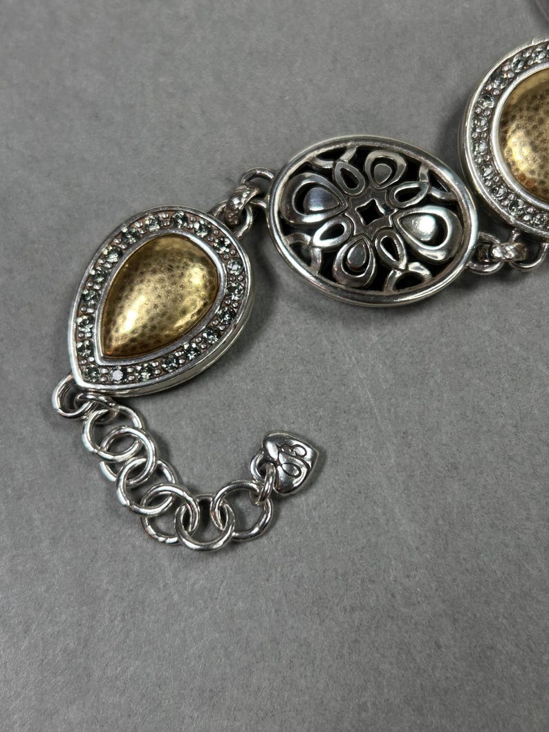 以下が含まれることがあります： 3つの装飾的なリンクを備えたシルバーカラーのブレスレット。リンクは、涙の形、花のパターン、円など、金と銀のアクセントがあります。ブレスレットは、小さな丸いリンクのチェーンで接続されています。