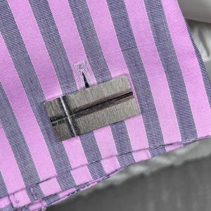 May include: Close-up of a striped dress shirt in pink and navy blue. A rectangular, silver-toned cufflink with a dark line detail is fastened to the shirt cuff. The shirt's fabric has vertical stripes.