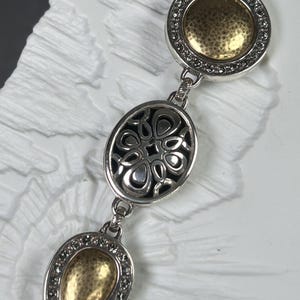 以下が含まれることがあります： ゴールドカラーのアクセントが付いたシルバーカラーのブレスレット。ブレスレットには、ゴールドの中央と小さな透明な石のリングが付いた丸いチャーム、透かし彫りのデザインの楕円形のチャーム、涙の形をしたチャームの3つの主要な装飾要素があります。