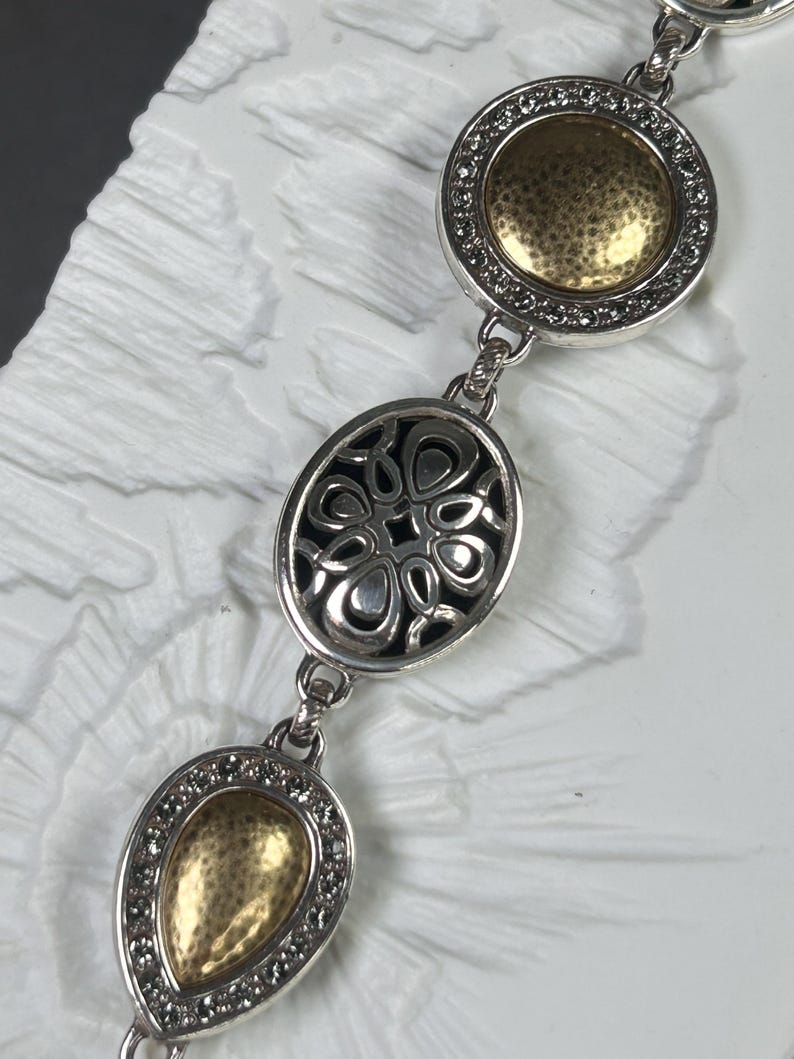 以下が含まれることがあります： ゴールドカラーのアクセントが付いたシルバーカラーのブレスレット。ブレスレットには、ゴールドの中央と小さな透明な石のリングが付いた丸いチャーム、透かし彫りのデザインの楕円形のチャーム、涙の形をしたチャームの3つの主要な装飾要素があります。