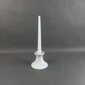 May include: A white ceramic candlestick holder with a textured, floral design base, holding a white taper candle. The candle is unlit. The candlestick holder is a decorative home accent.