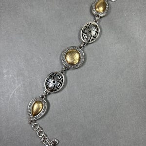 以下が含まれることがあります： ゴールドアクセントのシルバーブレスレット。ブレスレットは、涙型や楕円形など、一連の装飾的なリンクが特徴です。一部のリンクはテクスチャ加工されたゴールドの中央部を持ち、他のリンクは透かし彫りのデザインです。ロブスタークラスプ留めです。