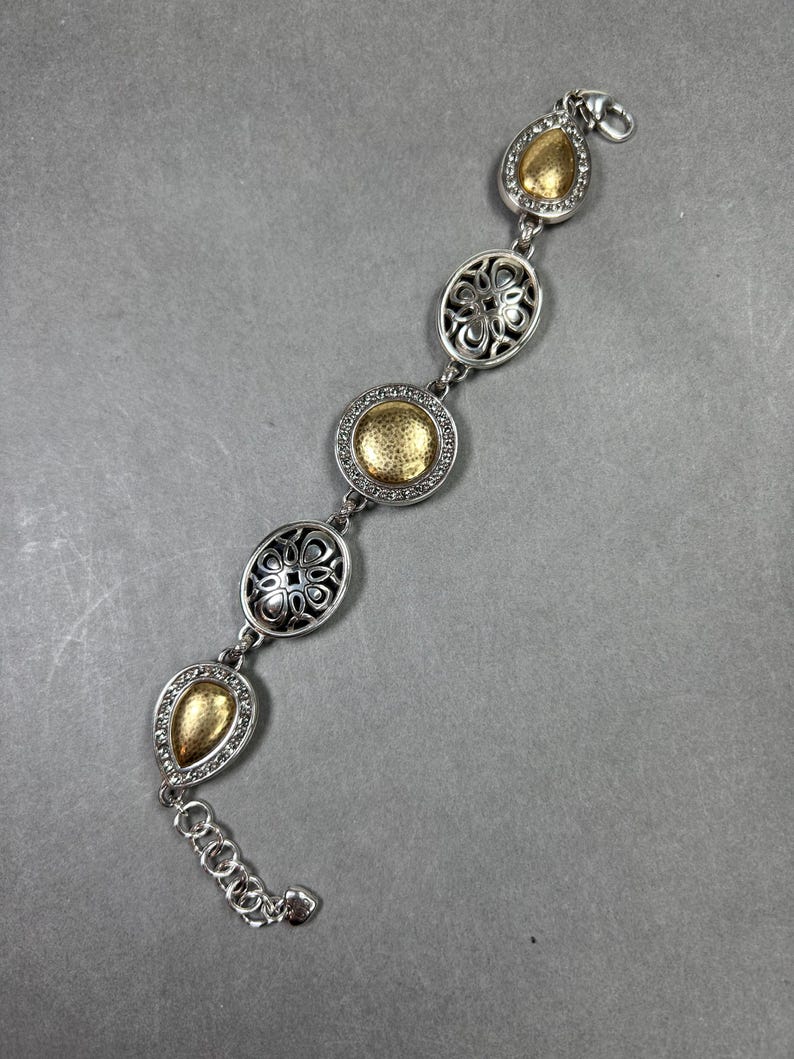 以下が含まれることがあります： ゴールドアクセントのシルバーブレスレット。ブレスレットは、涙型や楕円形など、一連の装飾的なリンクが特徴です。一部のリンクはテクスチャ加工されたゴールドの中央部を持ち、他のリンクは透かし彫りのデザインです。ロブスタークラスプ留めです。