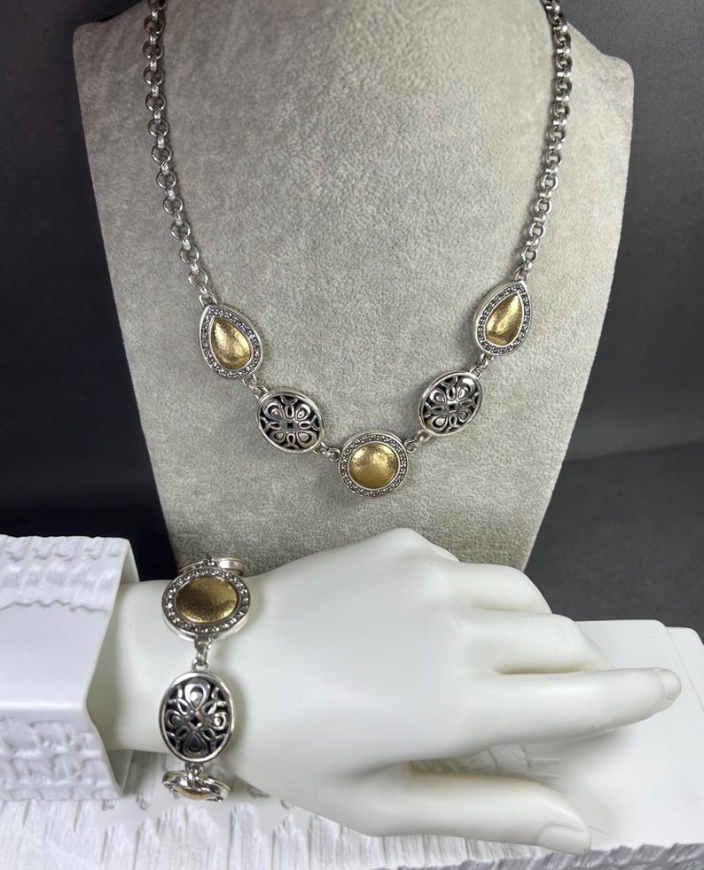 Peut inclure: Ensemble collier et bracelet argent&eacute;. Le collier pr&eacute;sente des accents dor&eacute;s en forme de goutte et d'ovale, ainsi que des motifs circulaires en filigrane. Le bracelet reprend le design du collier, avec des d&eacute;tails dor&eacute;s et en filigrane. Les bijoux sont pr&eacute;sent&eacute;s sur un mannequin.