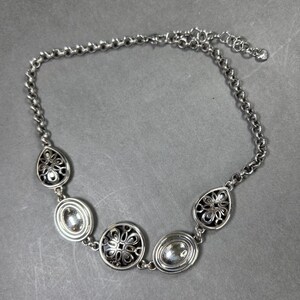 May include: Silver necklace with decorative pendants. The chain-link design features a lobster clasp. The pendants include oval and teardrop shapes with floral patterns and clear stone accents. The necklace is approximately 40 cm in length.