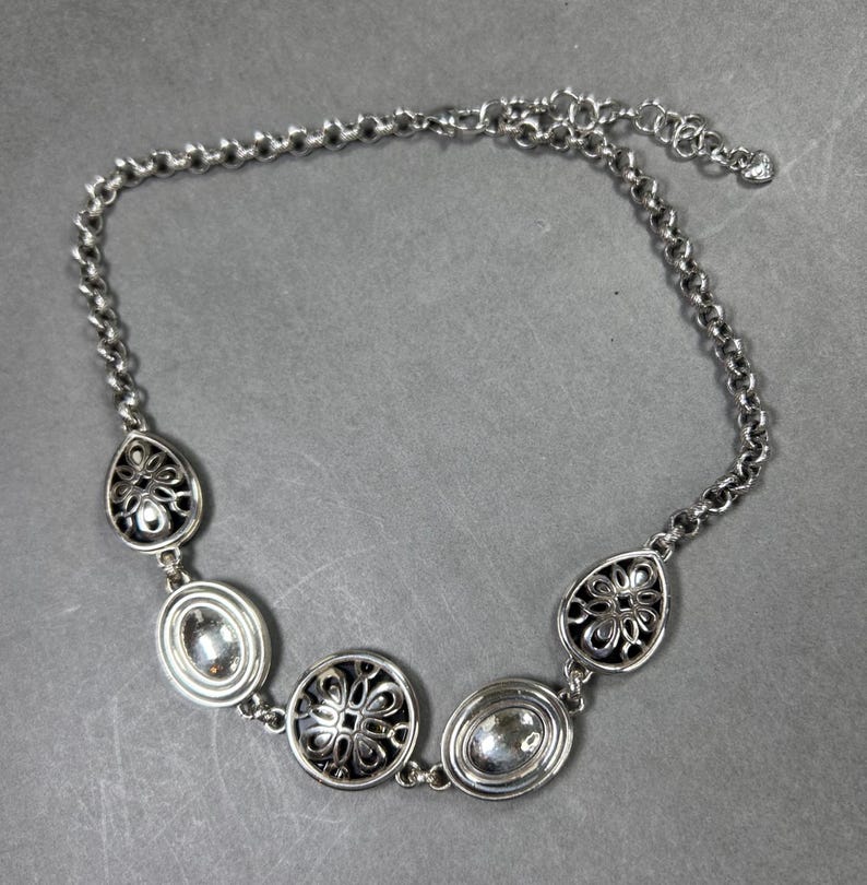 May include: Silver necklace with decorative pendants. The chain-link design features a lobster clasp. The pendants include oval and teardrop shapes with floral patterns and clear stone accents. The necklace is approximately 40 cm in length.