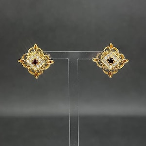 Puede incluir: Un par de pendientes de botón dorados. Cada pendiente presenta un diseño cuadrado con una piedra central de color rojo oscuro rodeada de piedras claras más pequeñas. Los pendientes tienen un borde ornamentado de estilo filigrana con pequeños detalles de color ámbar.