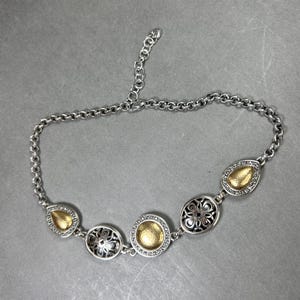 May include: Silver necklace with gold-coloured detailing. The necklace has a chain and decorative elements in various shapes, including teardrops and circles. The gold elements have a textured appearance, and the necklace has an adjustable length.