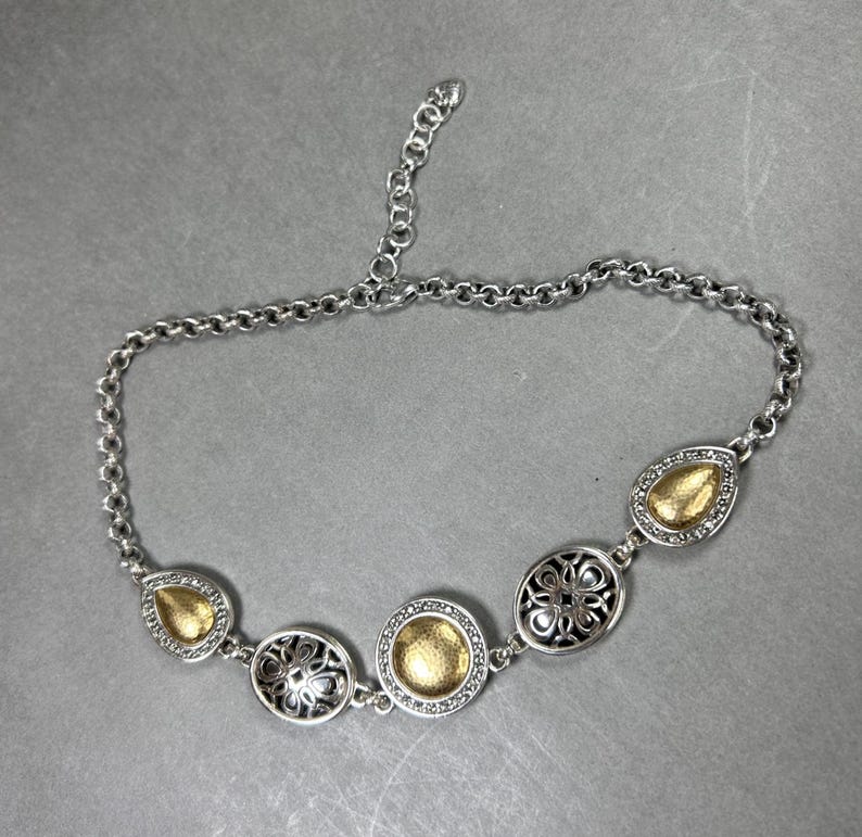 May include: Silver necklace with gold-coloured detailing. The necklace has a chain and decorative elements in various shapes, including teardrops and circles. The gold elements have a textured appearance, and the necklace has an adjustable length.