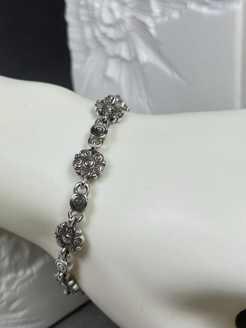 May include: Silver-toned bracelet featuring a series of floral and circular links. The bracelet is composed of multiple decorative flower-shaped elements, connected by smaller circular links. The design has an antique or vintage aesthetic.