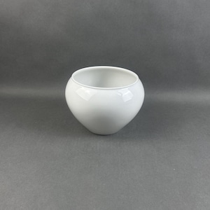 May include: A small, white, ceramic bowl with a rounded shape. The bowl has a smooth, glossy finish and a wide opening. The bowl is empty and sits on a gray surface. The bowl is likely used for serving food or as a decorative item.