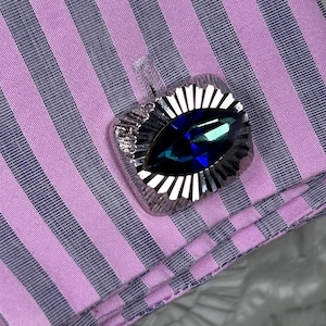 May include: A silver cufflink featuring a dark blue, teardrop-shaped gemstone. The cufflink is set against a pink and grey striped fabric. The design includes a sunburst pattern around the gemstone, creating a striking visual effect. The fabric is a striped pattern.