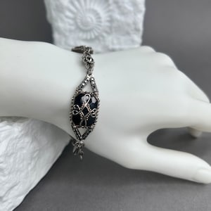 Könnte beinhalten: Silberfarbenes Armband mit einem großen, dunkelblauen Edelstein mit aufwendigem Filigranmuster. Das Armband hat eine zarte Kette und ist mit kleinen, funkelnden Akzenten verziert. Das Armband wird auf einer weißen Hand präsentiert.