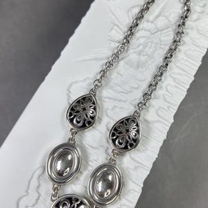May include: Silver necklace with a chain and decorative pendants. The necklace features oval and teardrop-shaped pendants with intricate black and silver floral designs. The chain is adjustable.