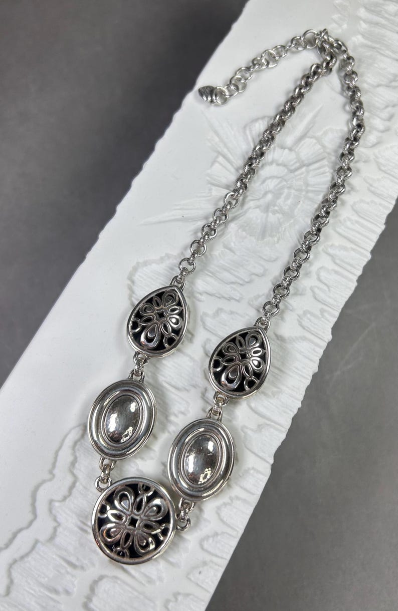 May include: Silver necklace with a chain and decorative pendants. The necklace features oval and teardrop-shaped pendants with intricate black and silver floral designs. The chain is adjustable.
