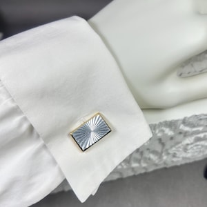 May include: A close-up of a white dress shirt cuff with a rectangular silver and gold cufflink. The cufflink has a silver, sunburst design within a gold frame. The cuff is on a white surface.