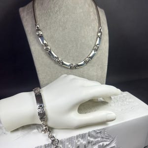 Peut inclure: Un ensemble collier et bracelet argent&eacute;. Le collier pr&eacute;sente un cordon marron avec des accents rectangulaires et ronds argent&eacute;s. Le bracelet a une plaque rectangulaire avec des &eacute;l&eacute;ments d&eacute;coratifs et une cha&icirc;ne avec un charme. Les bijoux sont expos&eacute;s sur un mannequin gris et une main blanche.