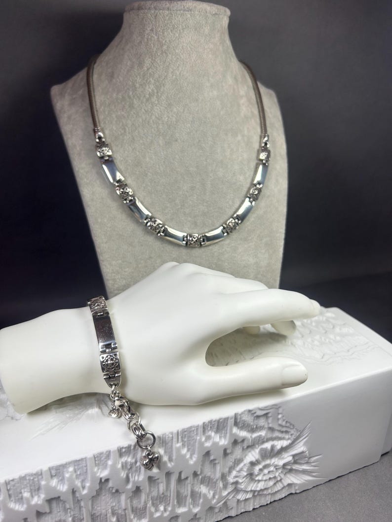 Peut inclure: Un ensemble collier et bracelet argent&eacute;. Le collier pr&eacute;sente un cordon marron avec des accents rectangulaires et ronds argent&eacute;s. Le bracelet a une plaque rectangulaire avec des &eacute;l&eacute;ments d&eacute;coratifs et une cha&icirc;ne avec un charme. Les bijoux sont expos&eacute;s sur un mannequin gris et une main blanche.