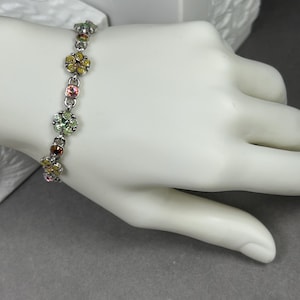 May include: A silver bracelet with colorful flower-shaped charms. Each charm features a cluster of small, round gemstones in shades of yellow, pink, green, and brown. The bracelet is displayed on a white hand against a gray background.