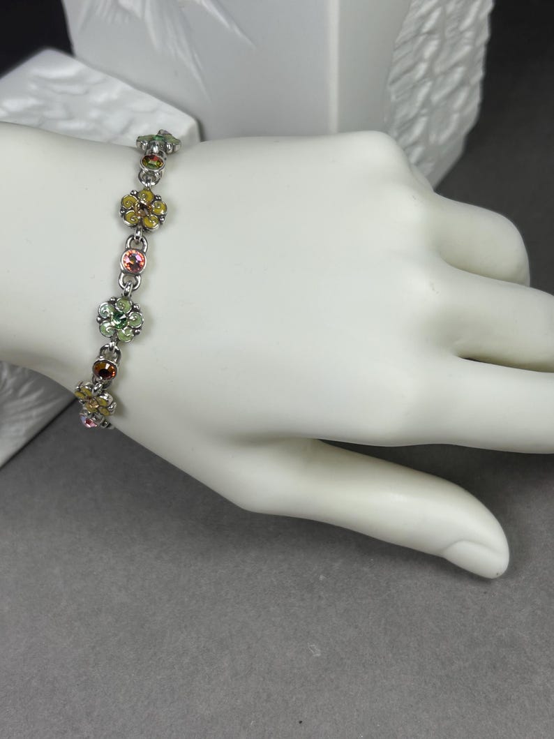 May include: A silver bracelet with colorful flower-shaped charms. Each charm features a cluster of small, round gemstones in shades of yellow, pink, green, and brown. The bracelet is displayed on a white hand against a gray background.