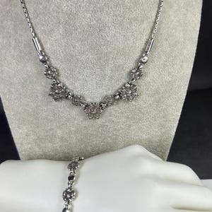 May include: A silver-toned necklace and bracelet set. The necklace features a delicate chain with floral-shaped pendants. The bracelet mirrors the design with similar floral accents. Both pieces are displayed on a mannequin.