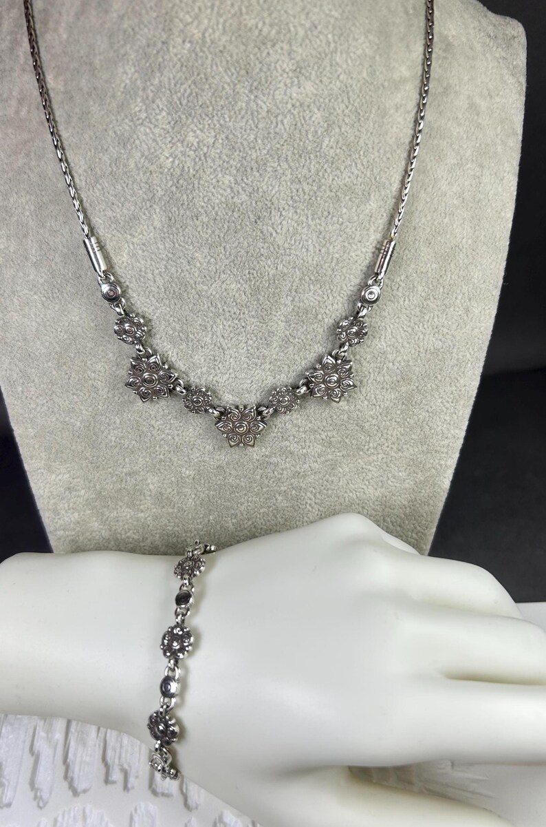 May include: A silver-toned necklace and bracelet set. The necklace features a delicate chain with floral-shaped pendants. The bracelet mirrors the design with similar floral accents. Both pieces are displayed on a mannequin.