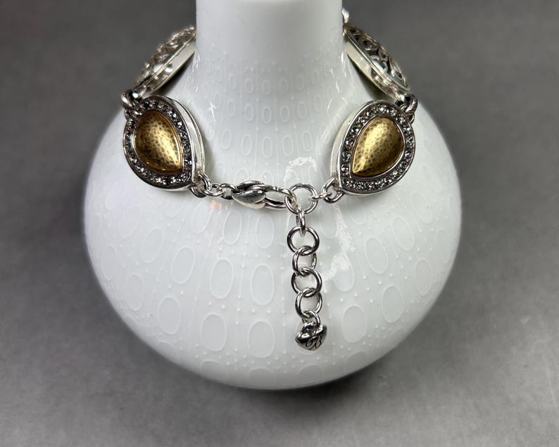 以下が含まれることがあります： ゴールドカラーのしずく型アクセントが付いたシルバーカラーのブレスレット。各しずくは、小さな透明なクリスタルで縁取られています。ブレスレットには、サイズ調整用のチェーンエクステンションとロブスタークラスプが付いています。