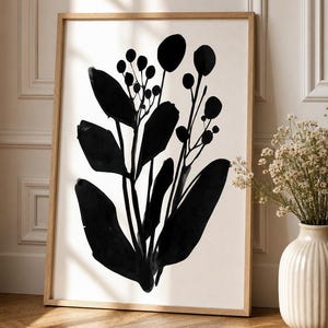 Op de afbeelding: Een rechthoekige wandkunstprint in een lichthouten lijst met een gedurfde zwarte inktillustratie van een botanische plant met langwerpige bladeren en kleine ronde bessen op een effen witte achtergrond.