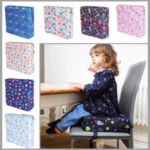 May include: A set of colourful booster seats for children. The seats feature various patterns, including dinosaurs, animals, and outer space. The seats are designed to help children reach the table and sit comfortably.