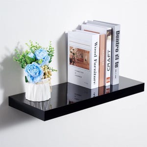 May include: A black floating shelf displaying books and a floral arrangement. The shelf supports several books with white and tan covers. A small vase with blue flowers and greenery is on the left side of the shelf.