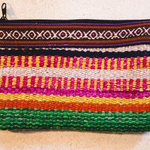 Könnte beinhalten: Eine rechteckige gewebte Clutch mit Reißverschluss und Handschlaufe. Die Clutch hat horizontale Streifen in verschiedenen Farben, darunter Grün, Orange, Gelb, Pink, Weiß, Schwarz und Braun. Der obere Bereich hat ein geometrisches Muster.