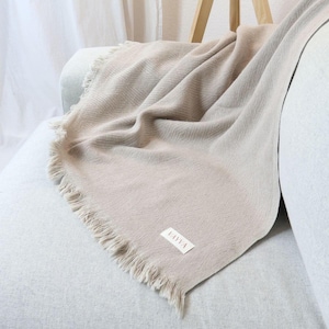 Könnte beinhalten: Eine beige Wolldecke mit Fischgrätenmuster und Fransen an den Rändern. Die Decke liegt auf einem hellgrauen Sofa und hat ein kleines "VAVA"-Etikett.