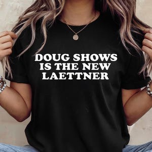 Könnte beinhalten: Schwarzes T-Shirt mit dem weißen Text "DOUG SHOWS IS THE NEW LAETTNER". Das Shirt hat einen Rundhalsausschnitt und kurze Ärmel. Das Model trägt eine goldene Halskette und Armbänder.
