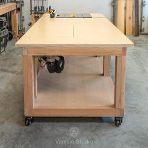 May include: A large wooden workbench featuring a light-coloured plywood top with an integrated table saw insert. The base is constructed from sturdy timber framing with a lower shelf and four black caster wheels for mobility in a workshop.