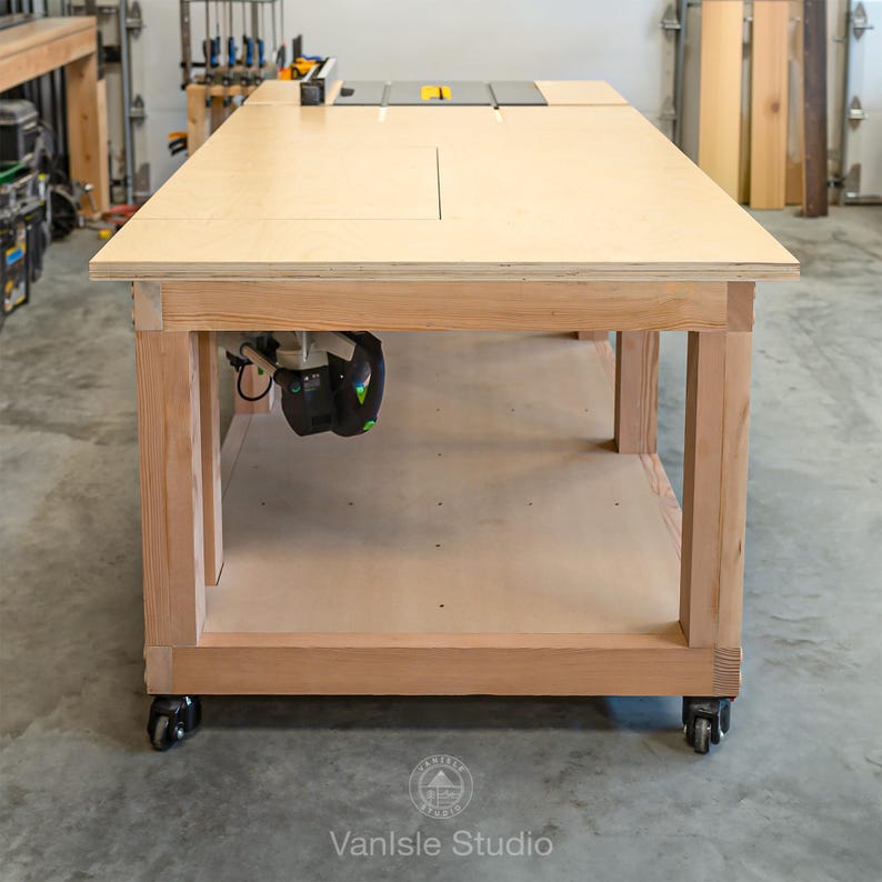 May include: A large wooden workbench featuring a light-coloured plywood top with an integrated table saw insert. The base is constructed from sturdy timber framing with a lower shelf and four black caster wheels for mobility in a workshop.