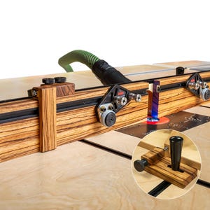 以下が含まれることがあります： ゼブラウッド製のルーターテーブルフェンス。黒いレールと調整可能なコンポーネントを備えています。緑色のホースと、同様のコンポーネントの小さなインセット画像も表示されます。テーブルは明るい色の木製の表面に置かれています。