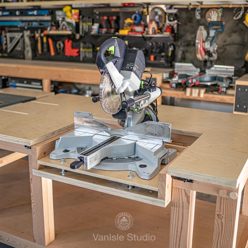 May include: A grey and black mitre saw with a circular blade, mounted on a light-coloured wooden workbench. The power tool features a green accent on the handle, a clear blade guard, and a metal base plate. The logo VanIsle Studio is visible.