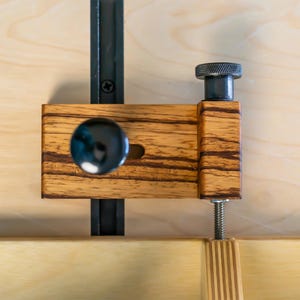 May include: A wooden woodworking clamp with a black metal rail and a black knob. The clamp has a striped wood grain pattern and a threaded rod for adjustment. The background is a light wood surface.