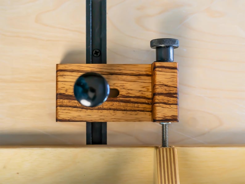 May include: A wooden woodworking clamp with a black metal rail and a black knob. The clamp has a striped wood grain pattern and a threaded rod for adjustment. The background is a light wood surface.