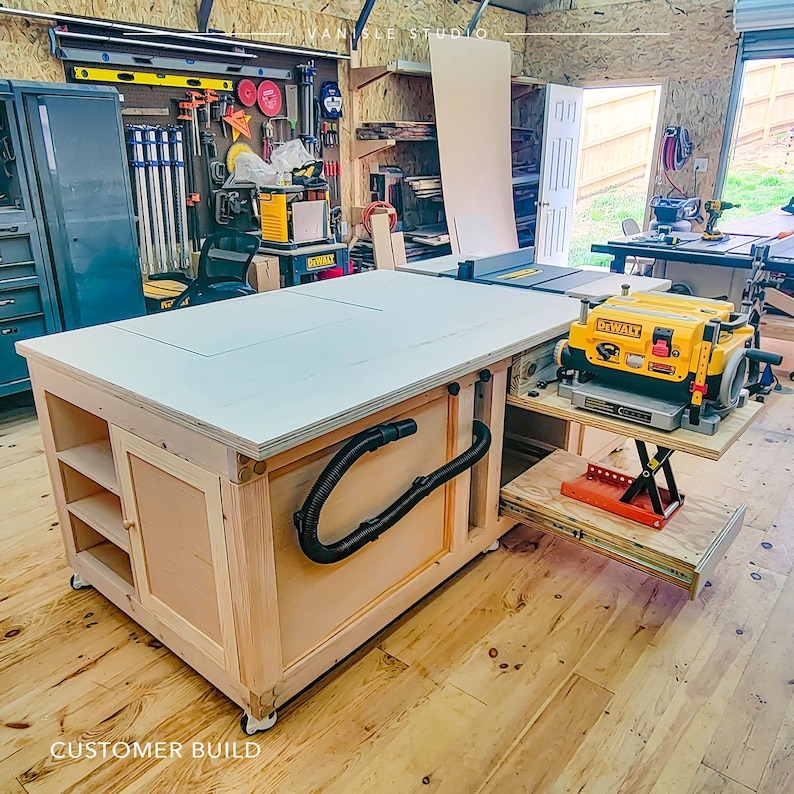 May include: A large, light-coloured wooden workbench on castors featuring an integrated table saw, open shelving, a cabinet door, and a black vacuum hose. A yellow and black DeWalt thickness planer sits on a retractable shelf with a red scissor lift.