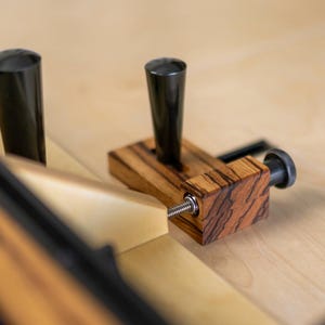 May include: A woodworking jig with a black handle and a wooden base with a zebrawood pattern. The jig has a black cylindrical post and a threaded screw for adjustment. The background is a light-colored wooden surface.