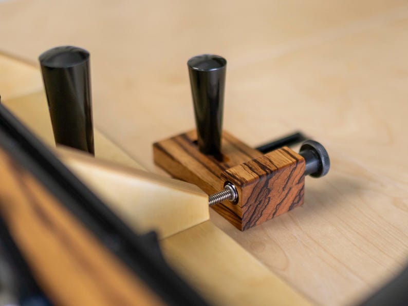 May include: A woodworking jig with a black handle and a wooden base with a zebrawood pattern. The jig has a black cylindrical post and a threaded screw for adjustment. The background is a light-colored wooden surface.
