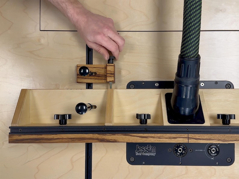 May include: A woodworking router table with a wooden fence, black knobs, and a dust collection hose. The table features a black base with the "JessEm Tool Company" logo. A hand is adjusting a part of the table.