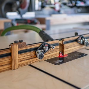 May include: A woodworking router table fence with a zebrawood finish. The fence has black hardware and adjustable components. The table is set up on a light-colored wooden surface.