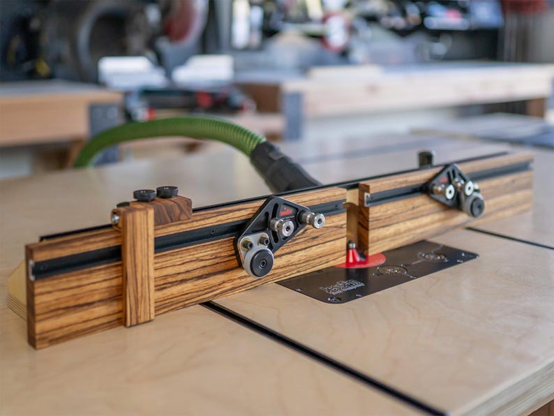 May include: A woodworking router table fence with a zebrawood finish. The fence has black hardware and adjustable components. The table is set up on a light-colored wooden surface.