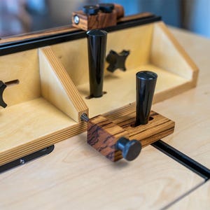 May include: A wooden router table with black knobs and handles. The table features a light-colored wood construction with darker wood accents and black metal components. The design includes adjustable elements and a sturdy build, suitable for woodworking.