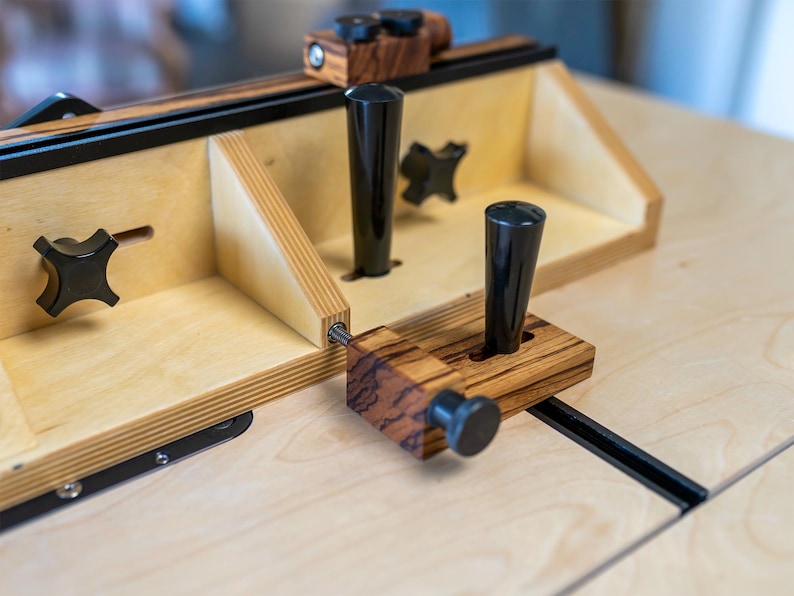 May include: A wooden router table with black knobs and handles. The table features a light-colored wood construction with darker wood accents and black metal components. The design includes adjustable elements and a sturdy build, suitable for woodworking.